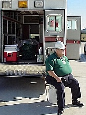 photo of KD5QXF, SK, sitting on back of old ambulance used as rehab supply van, holding radio in hand