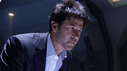 John Sheppard in suit coat and shirt, leaning on a table, showing his one tell at bluffing: the blep.