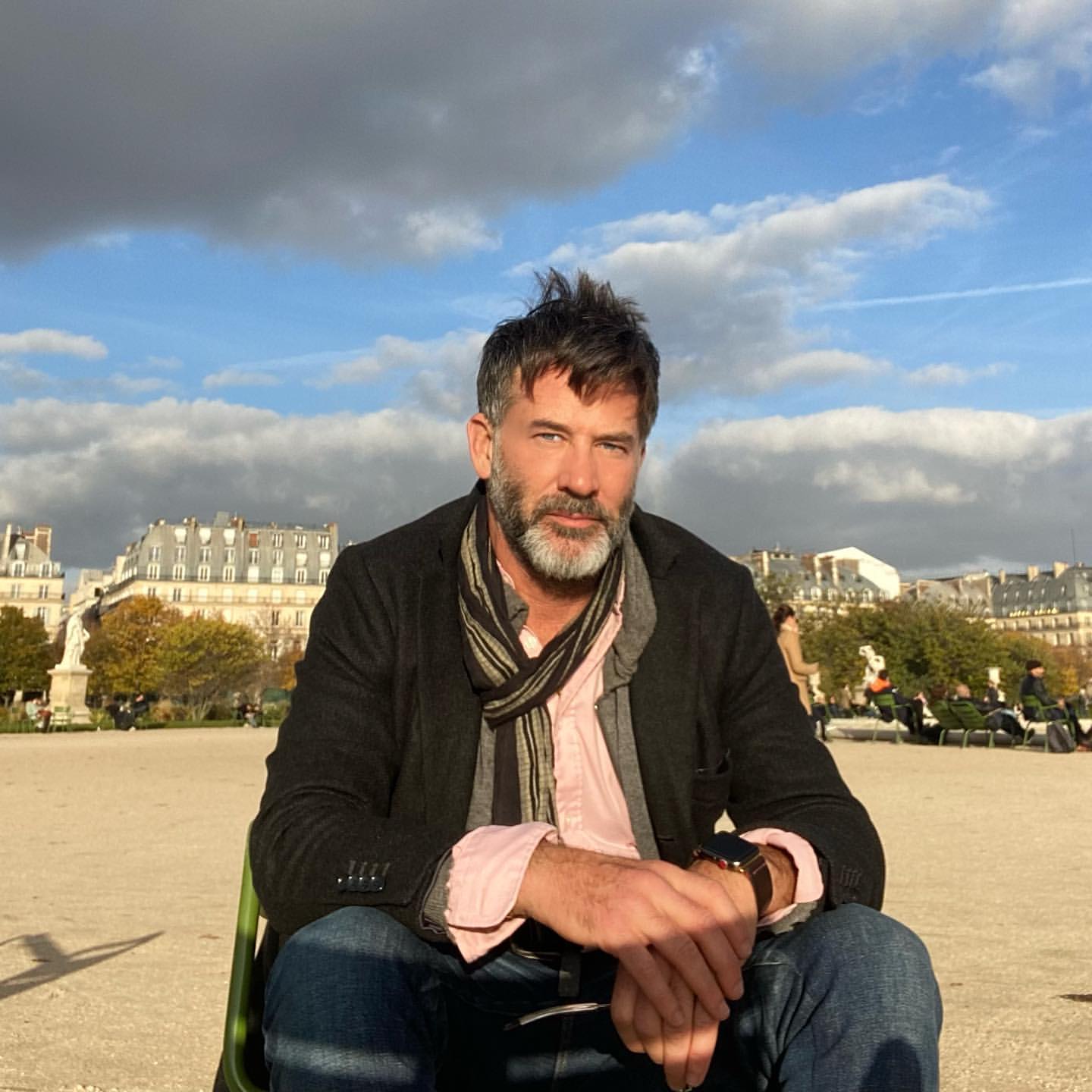 Joe with the Jardins de Louvre behind him. He is seated with one hand clasping the other wrist and wearing jeans and a black jacket over a pink button-down shirt, a gray and black scarf draped around his neck.