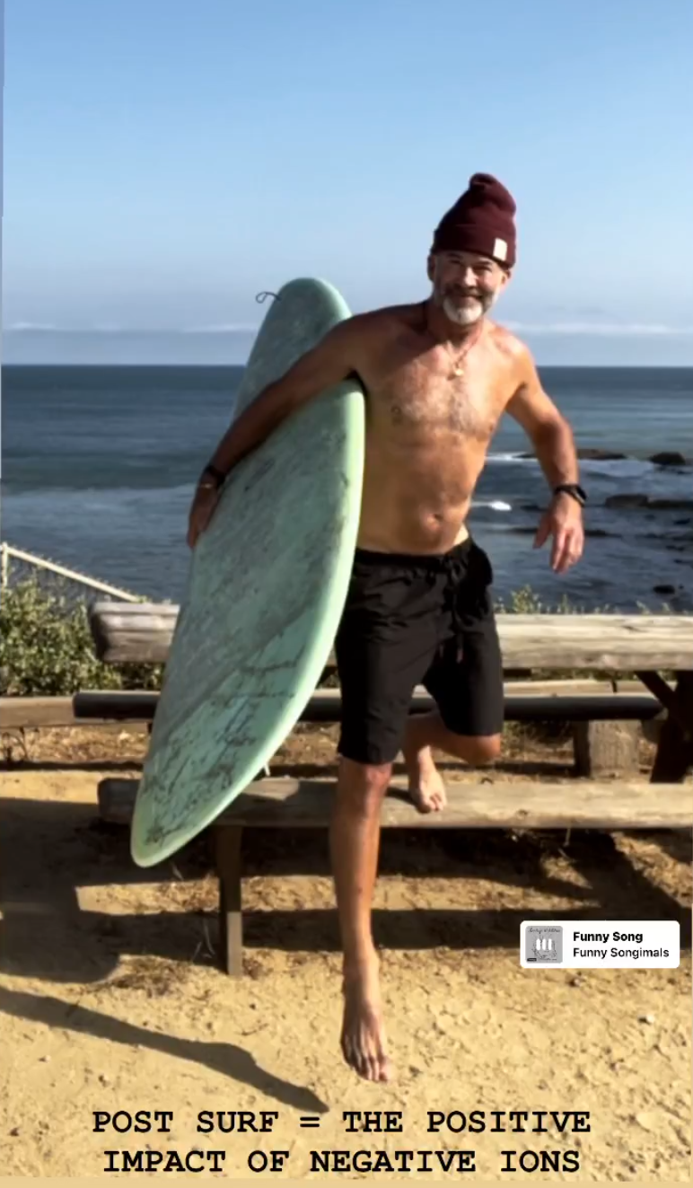 Joe Flanigan in black board shorts and a burgandy stocking cap holding a mint green surfboard, stepping down from a picnic table on the beach at Malibu. 