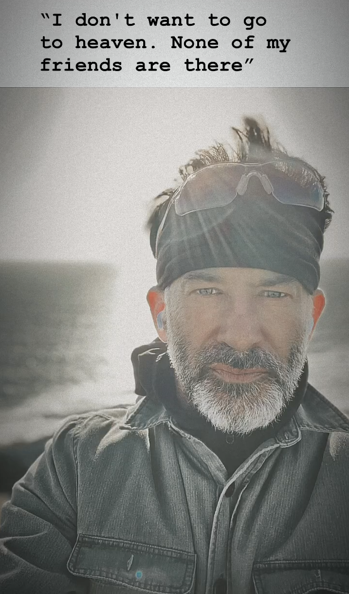 Joe Flanigan lit from behind in a black bandana, ski goggles, a black turtleneck and a gray jacket. His eyes glow green and are ringed with blue, and the caption reads, "I don't want to go to heaven. None of my friends are there."