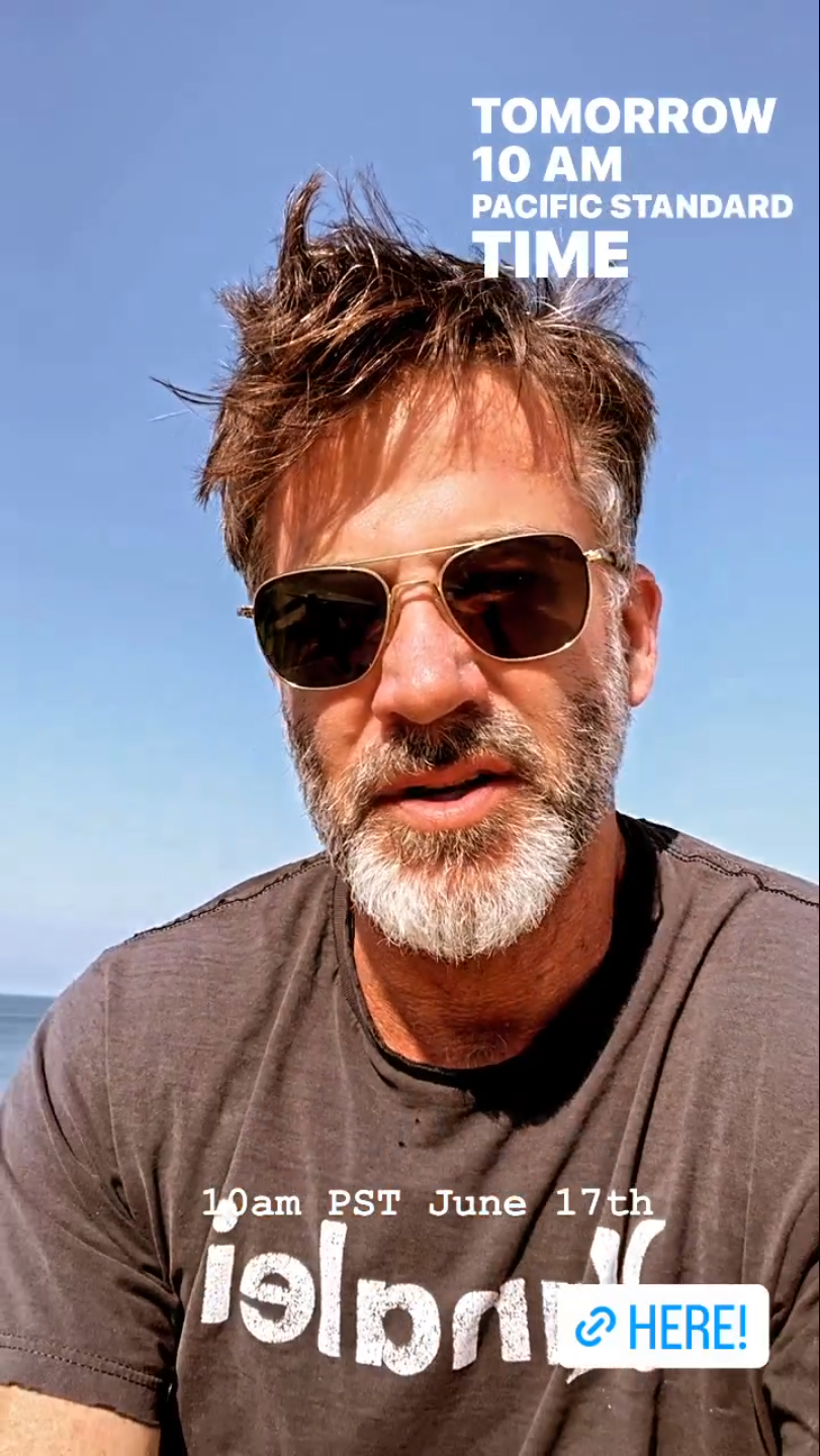 Joe Flanigan in a black t-shirt, his hair windswept. He's wearing aviators, the sun on his face and the ocean behind him.