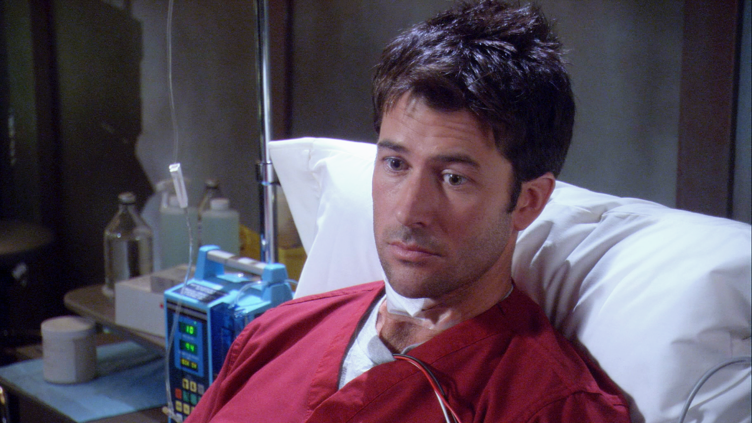 John in red scrubs with some electrical wires coming from his shirt. He's sitting in a hospital bed with monitoring equipment surrounding him and a plaintive expression on his face.