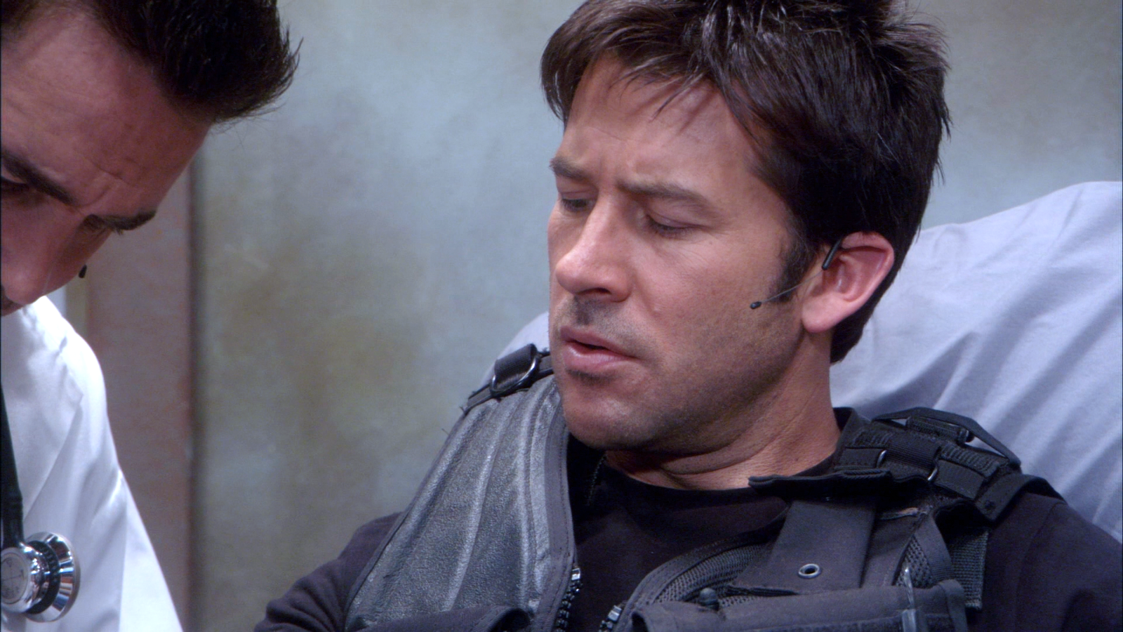 John in tac vest looking down at his arm with squirrely eyebrows while Carson Beckett does mysterious things to his arm.