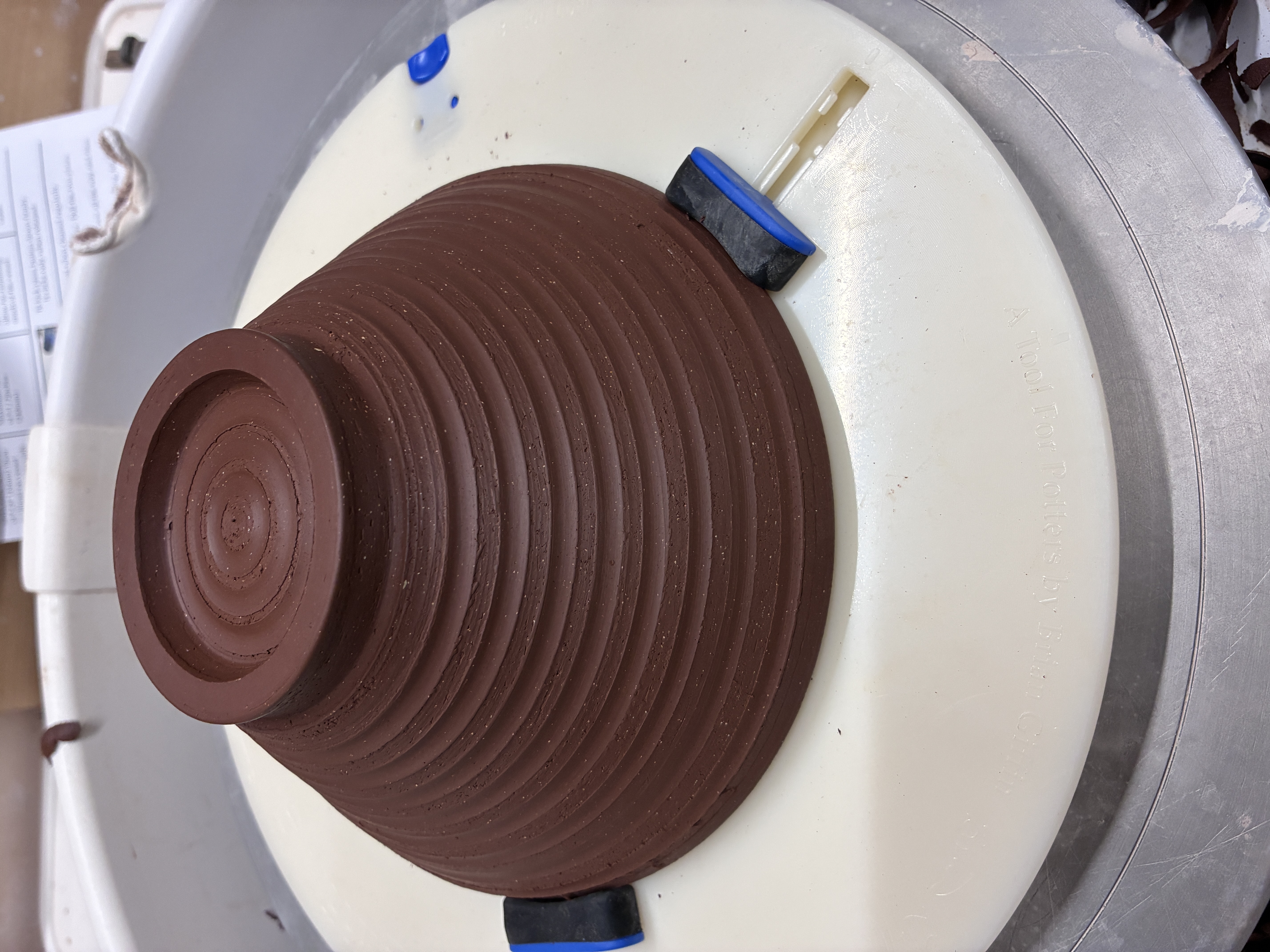 A dark brown clay bowl, upside down on a plastic trimming wheel, held in by three rubber coated clamps. The bowl has concentric rings on its sides and in its base.